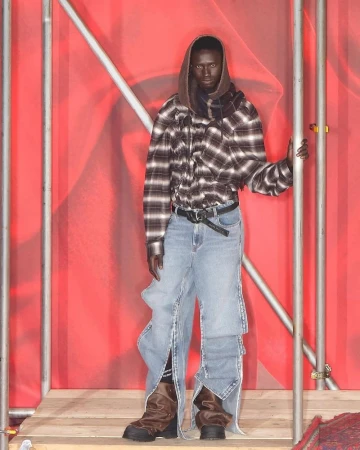 Runway fashion show: a model wears a hooded plaid shirt, baggy jeans, and boots, standing against a red backdrop with metal framing.