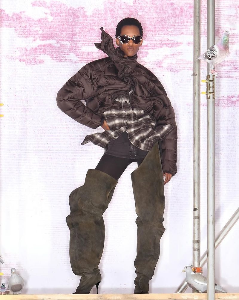 Fashion-forward woman poses in a brown puffer jacket, plaid shirt, and thigh-high olive green boots against a muted pink backdrop.