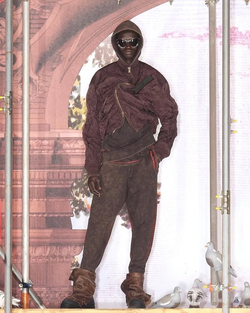 Runway model in brown jacket, hooded top, and pants stands on a platform with bird statues and ornate background details.