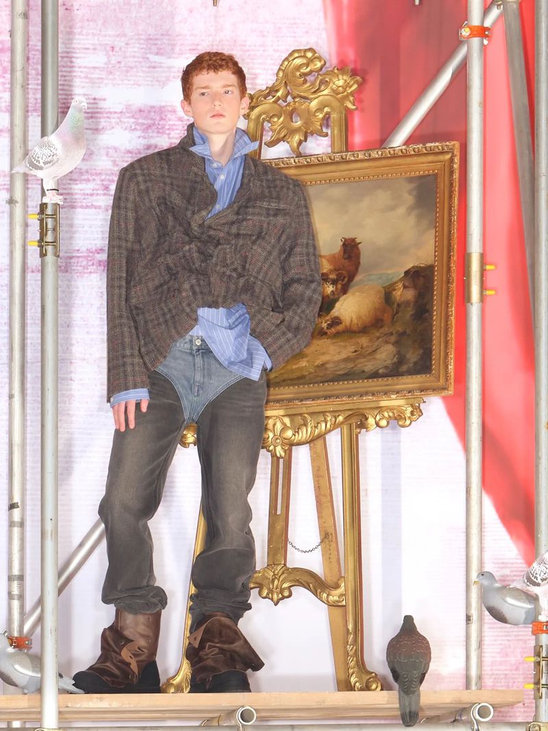A red-haired male model poses in a gray plaid jacket, jeans, and boots, standing in front of an antique painting and scaffolding.