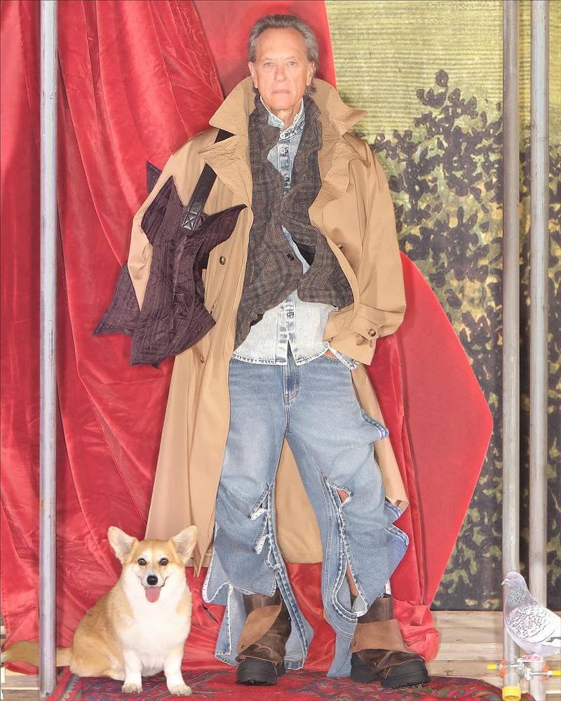 Mature adult male poses in a beige trench coat, ripped jeans, and brown boots next to a corgi in a staged indoor environment.