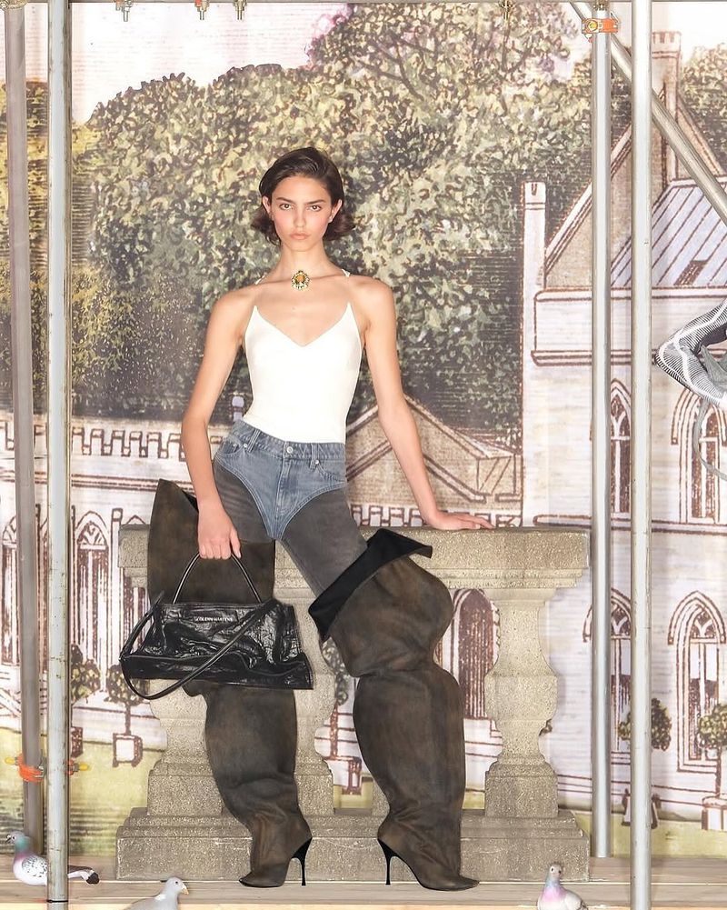 Fashion-forward shot of a woman in a white top, denim chaps, black bag, and oversized brown boots against a vintage mural backdrop.