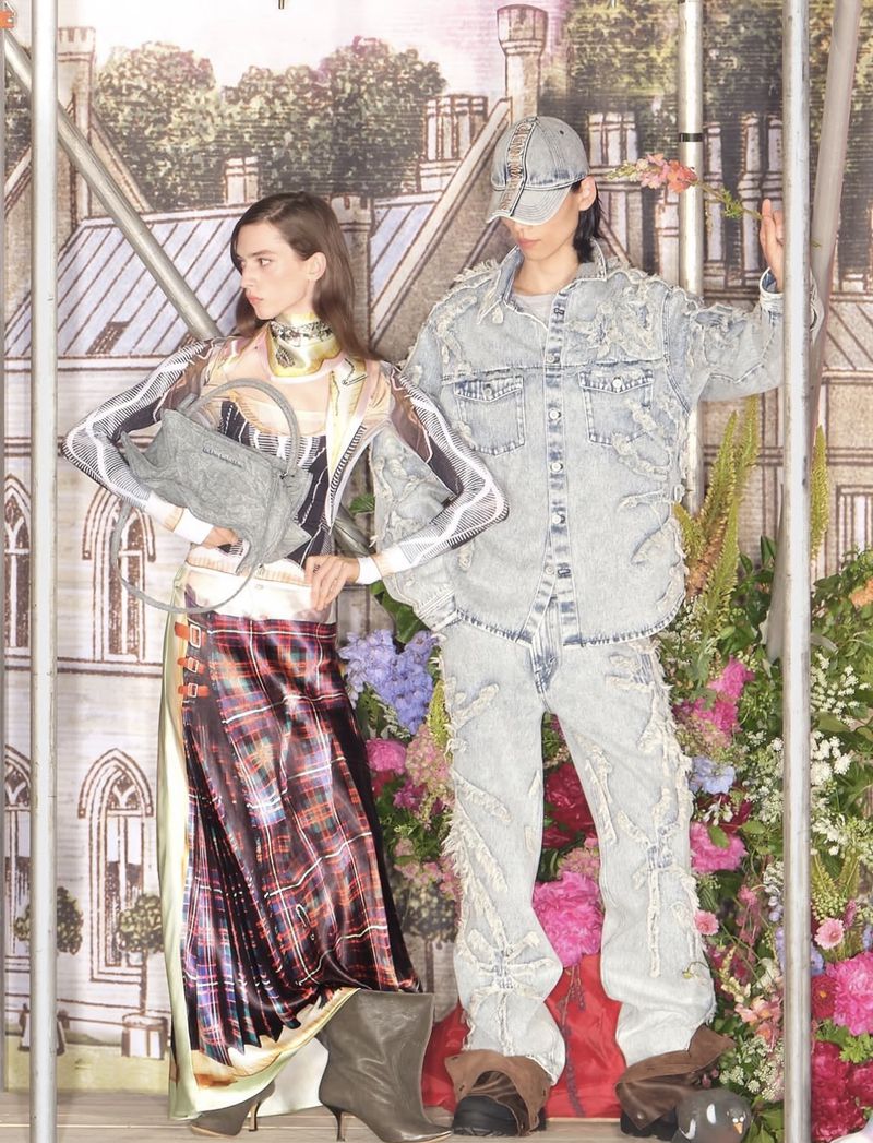 Two fashion models pose in front of a painted backdrop wearing deconstructed denim and plaid skirt with floral accents in a studio setting.