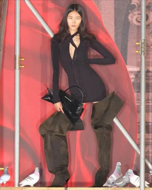 Model wearing a black dress and thigh-high boots holding a handbag against a red backdrop