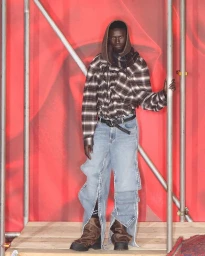 Black man wearing a plaid hooded shirt and split-leg denim jeans standing on a wooden platform against a red backdrop with metal scaffolding poles