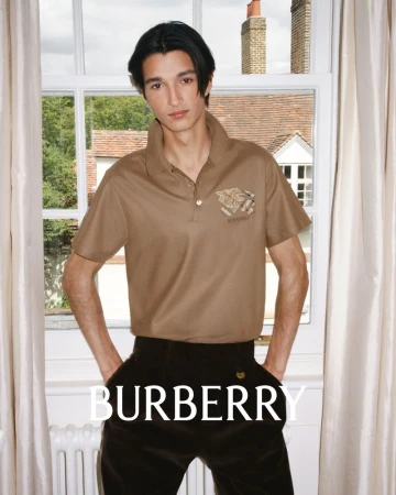 A young male model wearing a brown polo shirt stands indoors in front of a window featuring visible architectural details.