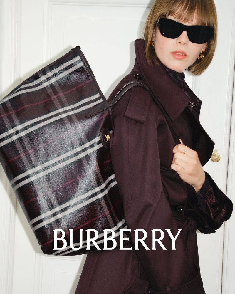 A woman with a burgundy trench coat and sunglasses carries a large Burberry plaid tote bag against a white paneled door.