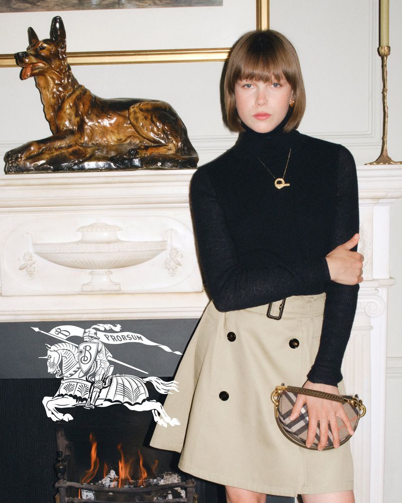 Woman in black turtleneck and trench skirt poses indoors near fireplace with Burberry handbag and ceramic dog sculpture on mantle.