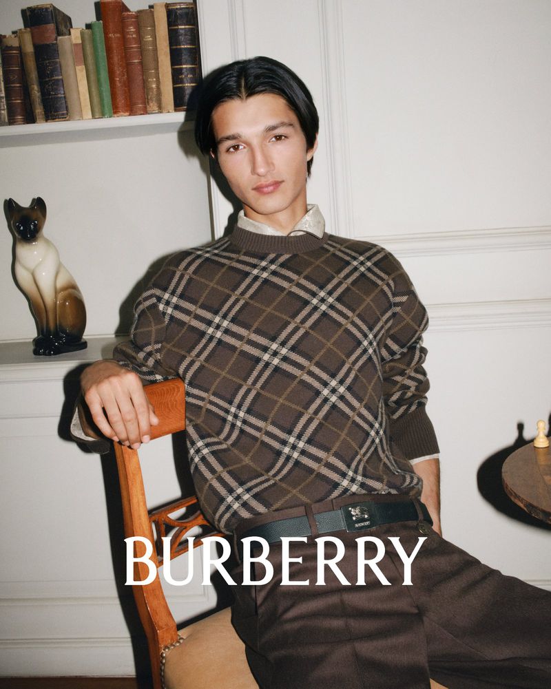 A stylish young man in a brown patterned sweater and brown pants poses in a vintage indoor setting, showcasing classic Burberry fashion.