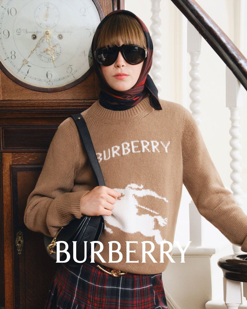 A young woman poses in a studio wearing a tan Burberry sweater with a horse graphic and a plaid skirt, along with a scarf and sunglasses.