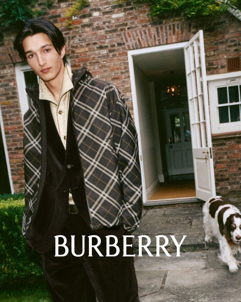 Man poses in plaid jacket and velvet suit with a Springer Spaniel dog in front of a brick house, promoting the Burberry brand.