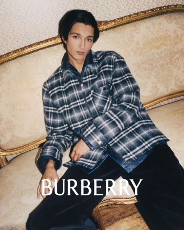 A fashionable adult male model poses indoors wearing a plaid jacket, black shirt and pants, sitting on a gold ornamented sofa.