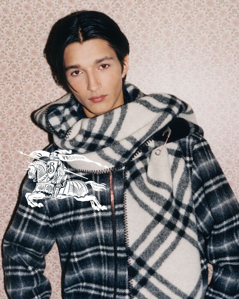 Male model in a hooded plaid jacket poses against a floral wallpaper backdrop in a studio environment. Fashion editorial photography.