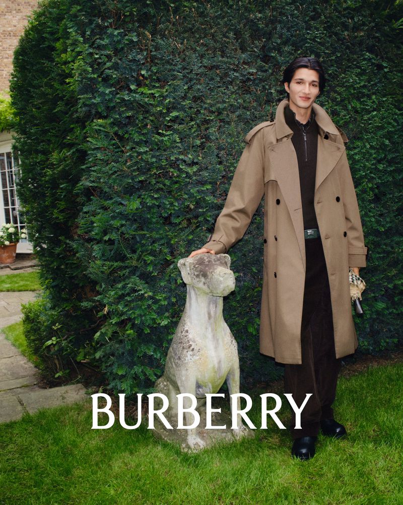An adult male model poses next to a dog statue, wearing a trench coat, in front of greenery, for a Burberry fashion campaign.