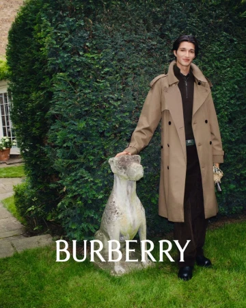 An adult male model poses next to a dog statue, wearing a trench coat, in front of greenery, for a Burberry fashion campaign.