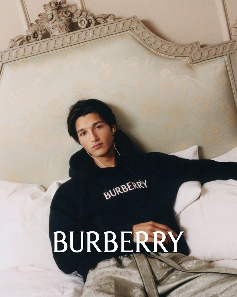 A relaxed, fashionable male model is posed on a bed wearing a black Burberry hoodie and patterned pants, showcasing casual luxury.