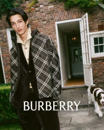 Man wearing plaid hooded jacket and dark trousers standing outdoors near a brick building with a dog