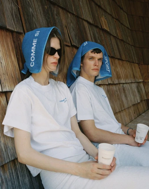 Two models wearing white T-shirts and blue head towels pose together outside with drinks, showcasing a casual and sporty aesthetic.