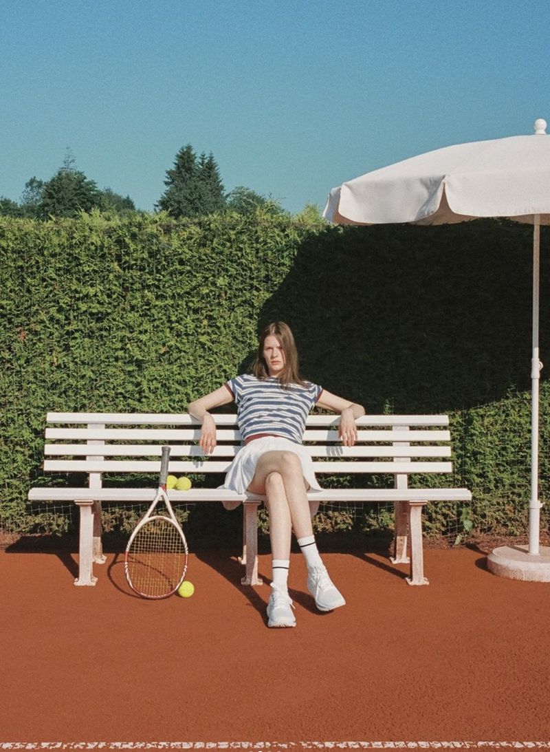 A young adult woman in a tennis outfit sits on a bench next to a tennis racket and balls, with a tennis court and hedge in the background.