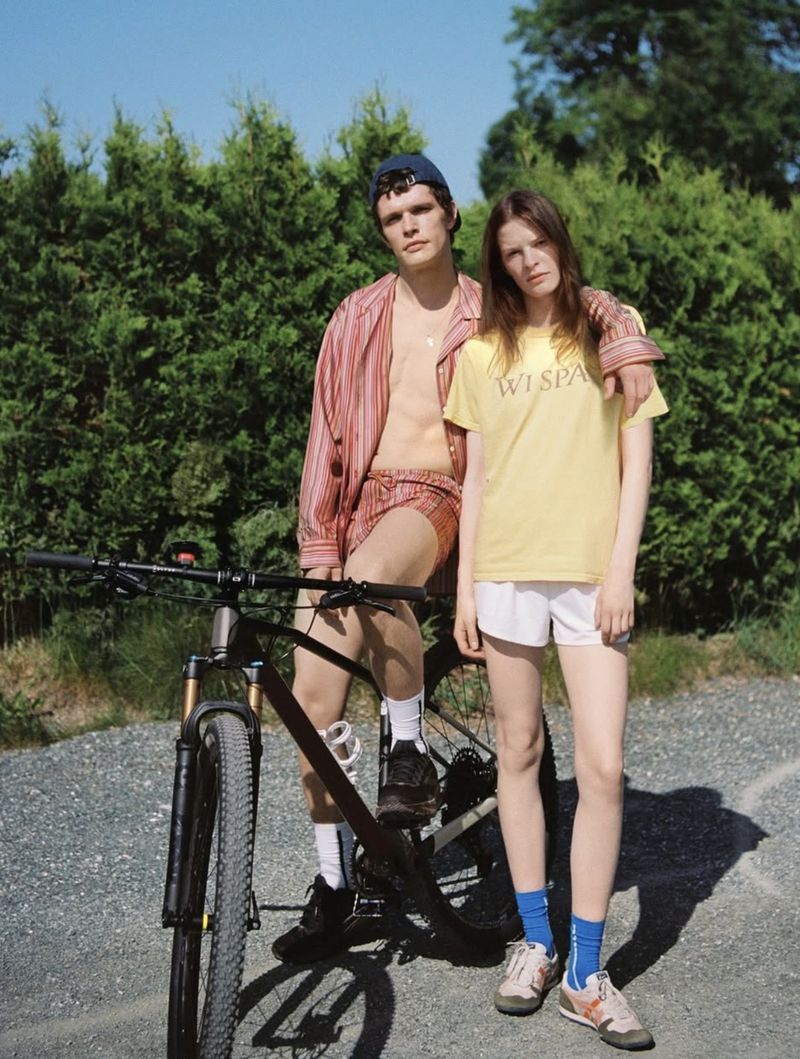 A couple wearing casual clothing poses next to a mountain bike outdoors surrounded by green foliage and a gravel path.