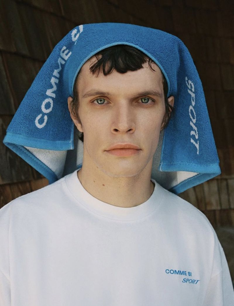 An androgynous adult model poses looking straight ahead in a white T-shirt and a bright blue graphic towel on their head indoors.