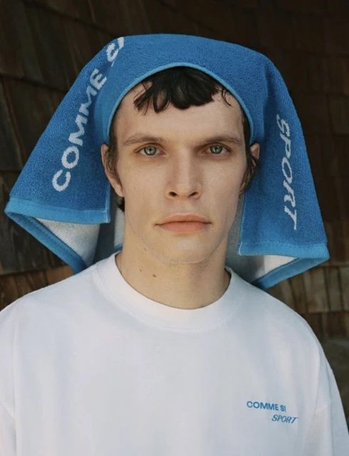 An androgynous adult model poses looking straight ahead in a white T-shirt and a bright blue graphic towel on their head indoors.
