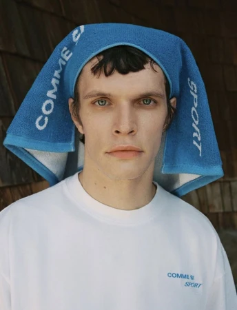 An androgynous adult model poses looking straight ahead in a white T-shirt and a bright blue graphic towel on their head indoors.