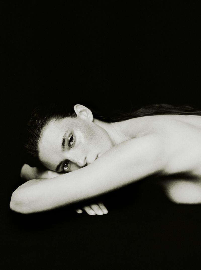 Monochrome portrait of an adult woman lying down with her arms crossed and looking at the camera against a dark backdrop.
