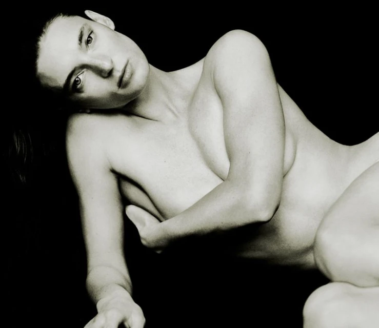 Portrait of a reclining adult woman in a black-and-white studio, gazing towards the camera with a hand covering her chest.