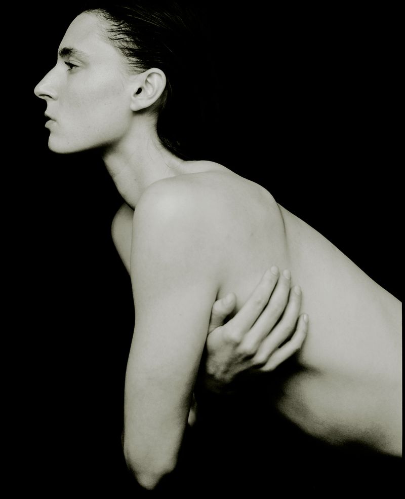 Monochromatic portrait of a slim, nude woman with wet hair and her arms crossed against a solid black background, minimalist and artistic.
