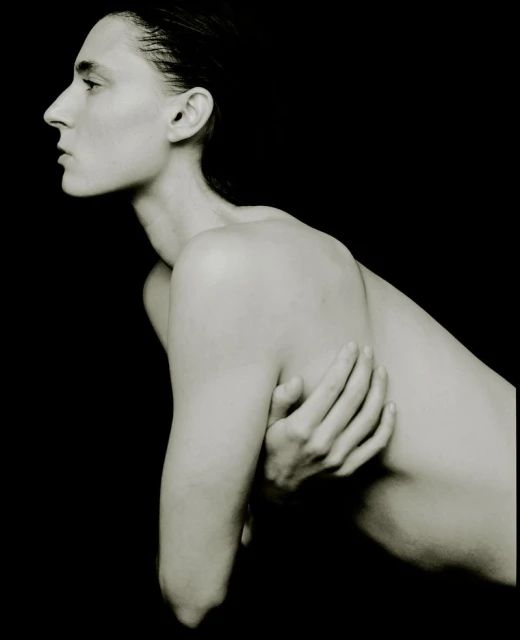 Monochromatic portrait of a slim, nude woman with wet hair and her arms crossed against a solid black background, minimalist and artistic.