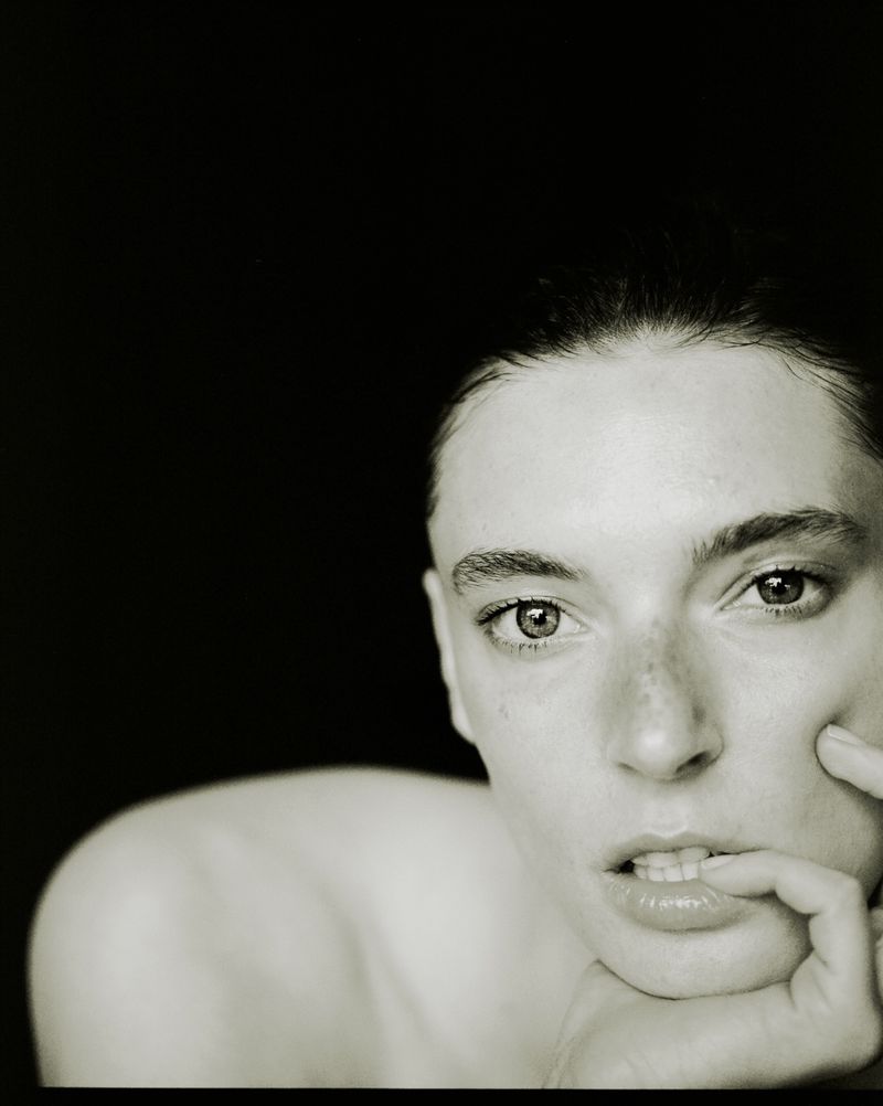 Close-up portrait of a fair-skinned young woman with dark hair and eyes, biting her lip against a dark background, shot in black and white.