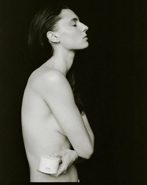 Monochrome studio shot: a young adult with slicked-back hair and closed eyes, holding a white cosmetic jar against a stark black background.