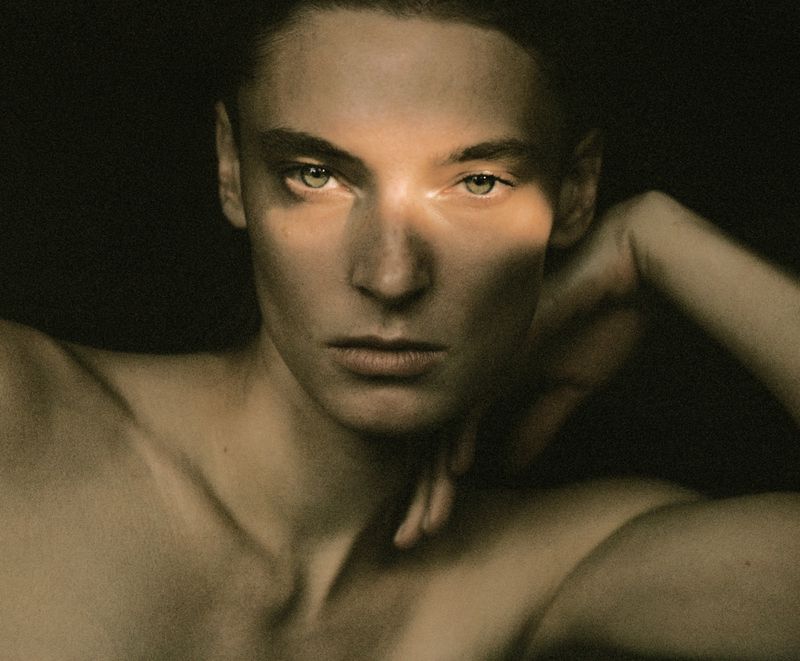 Close-up portrait of a model with dramatic lighting casting light bands across her face, raising one hand near her head.