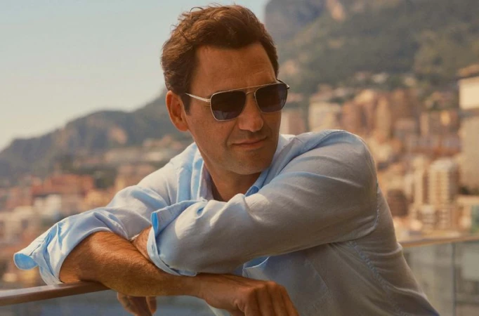 Adult male in sunglasses and light blue shirt leaning on a rail with a blurred background of buildings and mountains.