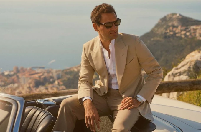 Adult male in a beige suit and sunglasses sits on a car near mountains and a body of water on a sunny day, looking off to the side.