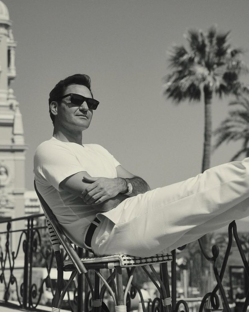 Monochrome shot of a man in sunglasses sitting in a chair outdoors, wearing all white, with a building and palm trees in the background.