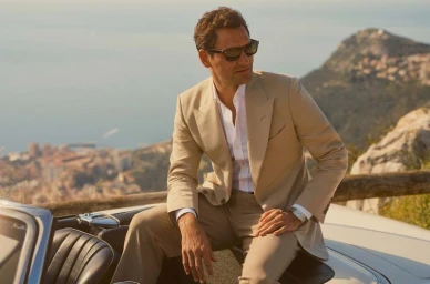 Roger Federer wearing Oliver Peoples sunglasses and a beige suit seated in a convertible car with a coastal background.