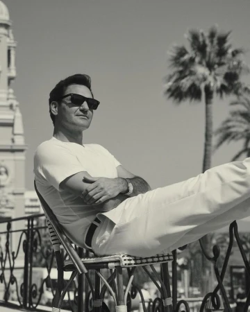 Roger Federer wearing sunglasses and white clothing, seated outdoors on a chair with feet propped on a railing next to a palm tree.