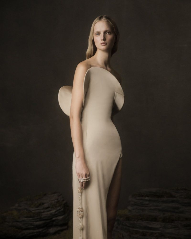 A woman in a beige gown poses in front of a dark background. She wears a beaded bracelet and ring. The dress has a large architectural detail.