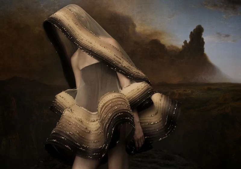 Woman in elaborate, layered dress with neutral tones and beaded accents poses in front of a dramatic landscape painting.