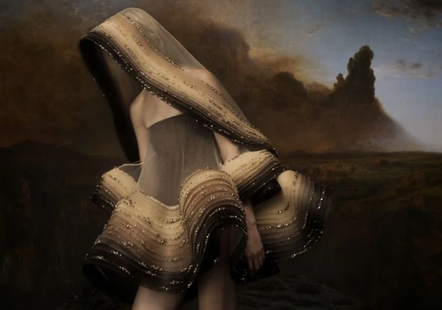 Woman in elaborate, layered dress with neutral tones and beaded accents poses in front of a dramatic landscape painting.