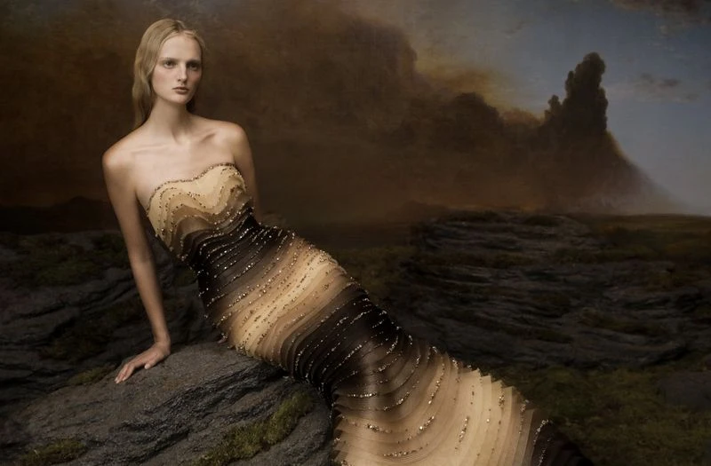 A fair-skinned woman in a beaded ombre gown poses on a rocky surface with a dramatic dark sky and landscape backdrop.