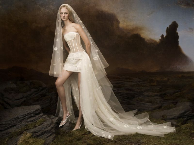 Model in white strapless corset dress and sheer veil posing on rocky terrain against dramatic stormy sky