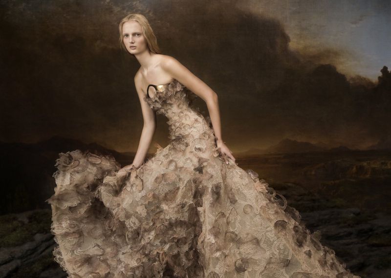 Fashionable adult female model poses in an elegant floral gown against a dark, painterly landscape background in a studio setting.
