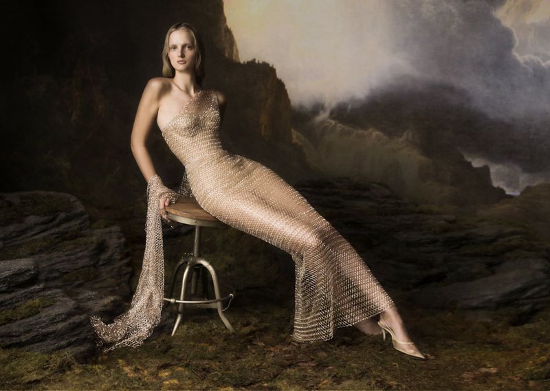 A fair-skinned woman poses in a full-length, gold-toned, chain-mail-style gown on a stool, set against a dramatic, painterly storm-cloud background.
