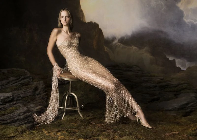 A fair-skinned woman poses in a full-length, gold-toned, chain-mail-style gown on a stool, set against a dramatic, painterly storm-cloud background.