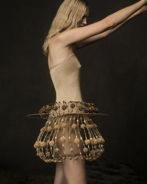 A slender adult woman is posing in a beige bodysuit and a pearl-embellished skirt against a dark studio backdrop, creating a fashion-forward visual.