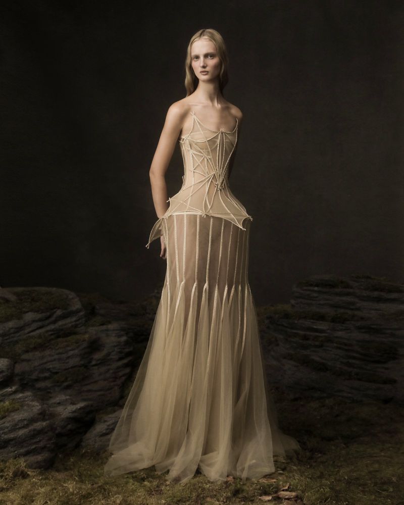 Blonde woman in a sheer beige gown with a web-like corset poses in a moody outdoor setting, with a neutral backdrop and rocky ground.