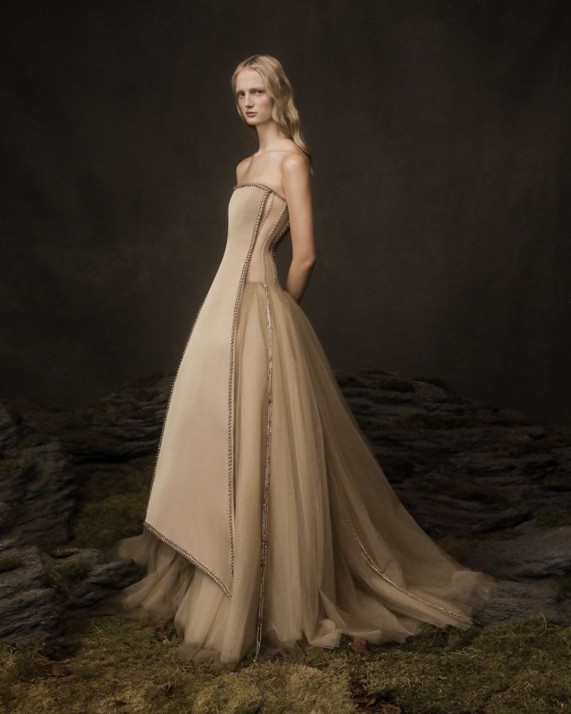 A woman in a beige strapless gown stands in a dark environment with rocks and moss in the foreground, in a fashion editorial shot.